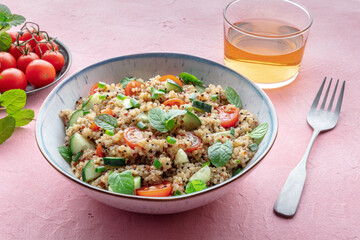 Quinoa tabbouleh salad in a bowl, a healthy dinner with tomatoes and mint, with a fork and a drink on a pink background