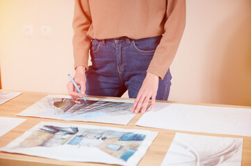 A young female illustrator carefully inspects her artwork on the table, evaluating each painted...