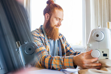 handsome redhaired man with long beard sews at a sewing machine at home studio
