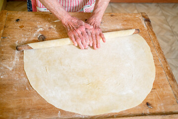 grandmother kneads dough, prepares noodles in the kitchen at home. Senior woman hands are rolling out dough in flour with a rolling pin in her home kitchen. 