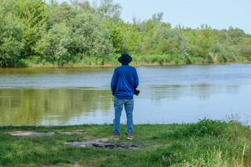 Fototapeta na wymiar Mężczyzna w kapeluszu ubrany w niebieski strój, stojacy tyłem, patrzący na rzekę 