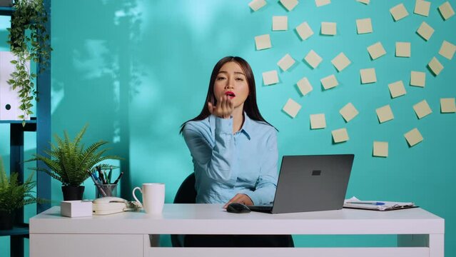 Frisky playful office clerk sending flirty funny air kisses while at work. Lively lighthearted businesswoman having fun in relaxed colourful workplace over blue studio background