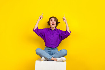 Full size photo of positive pretty girl sit podium look indicate fingers up empty space isolated on...