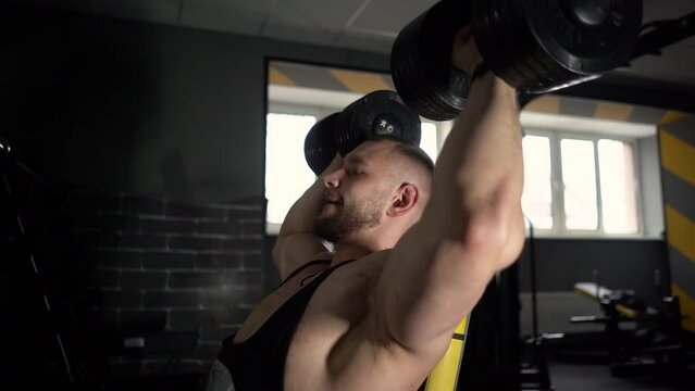 A strong male bodybuilder shakes his biceps in the gym with a barbell and dumbbells, at this time the muscles are tense