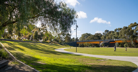 A public urban park with a large open outdoor space, well-maintained grass lawn, and a footbridge...