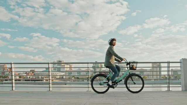 Long shot of modern Caucasian female office worker cycling along street on bank of the river in morning