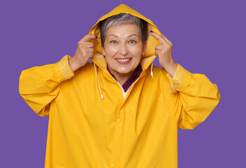Happy senior woman wearing yellow raincoat smile, poses on purple background. joyful expression showcase lively and positive spirit of elderly generation. Active lifestyle of a cheerful senior woman