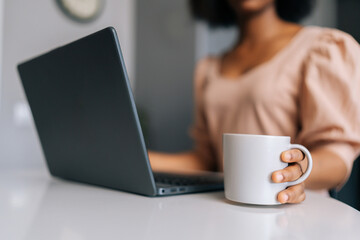 Close-up cropped shot of unrecognizable black female freelancer typing on laptop computer and holding cup with coffee or tea in morning sitting at table, working from remote home office workplace.