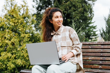 Cool cute young woman freelancer working on laptop outdoors. Smiling woman in casual clothes sitting with computer on park bench and looking away