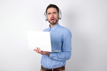 Shocked embarrassed businessman wearing blue t-shirt with headphones over white background keeps mouth widely opened. Hears unbelievable novelty stares in stupor