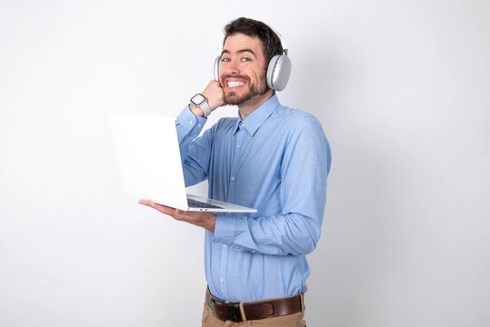 Happy businessman wearing blue t-shirt with headphones over white background keeps fists on cheeks smiles broadly and has positive expression being in good mood