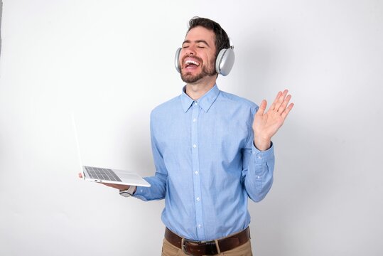 Overjoyed successful businessman wearing blue t-shirt with headphones over white background raises palm and closes eyes in joy being entertained by friends