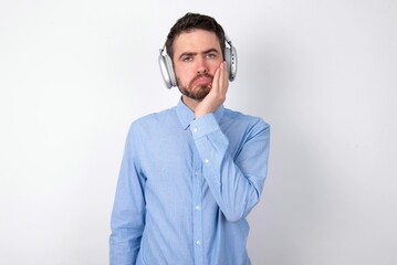 Sad lonely businessman wearing blue t-shirt with headphones over white background touches cheek with hand bites lower lip and gazes with displeasure. Bad emotions