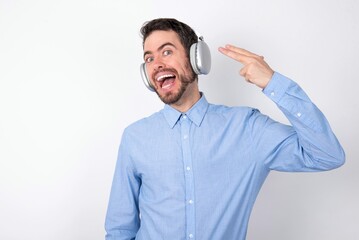 businessman wearing blue t-shirt with headphones over white background foolishness around shoots in temple with fingers makes suicide gesture. Funny model makes finger gun pistol