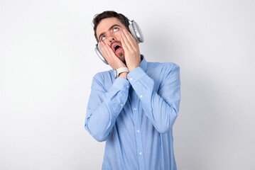 businessman wearing blue t-shirt with headphones over white background keeps hands on cheeks has bored displeased expression. Stressed hopeless model