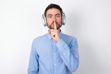 Surprised businessman wearing blue t-shirt with headphones over white background makes silence...