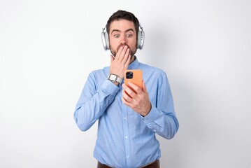 businessman wearing blue t-shirt with headphones over white background being deeply surprised, stares at smartphone display, reads shocking news on website, Omg, its horrible!