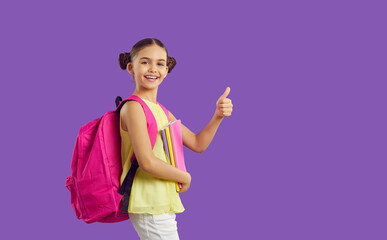 Smiling schoolgirl with backpack and books isolated on purple studio background show thumb up excited about school start. Happy girl pupil with notebooks recommend online study course. Education.