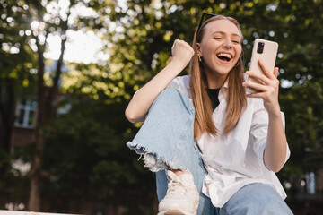 Smiling young girl sitting on ground browsing internet with cellphone gesturing like winner....