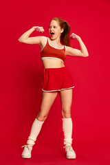 Charming red haired young girl wearing sportswear training with dumbbells with happy face on red background. Sport workout for slim body