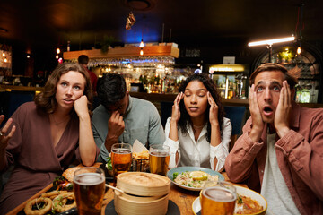 Frustrated young multiracial group of friends in casual clothing with dinner and drinks at restaurant