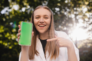 Happy attractive woman holding a mobile phone and point on green screen with message, news or recommendation. Mobile phone with a green screen for mockup. Happy smiling blonde woman.