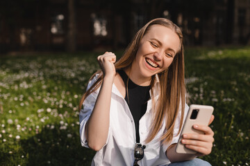 Smiling young girl sitting on ground browsing internet with cellphone gesturing like winner. Excited face expression, unable to believe that she bet played, win in lottery, can't believe in fortune.