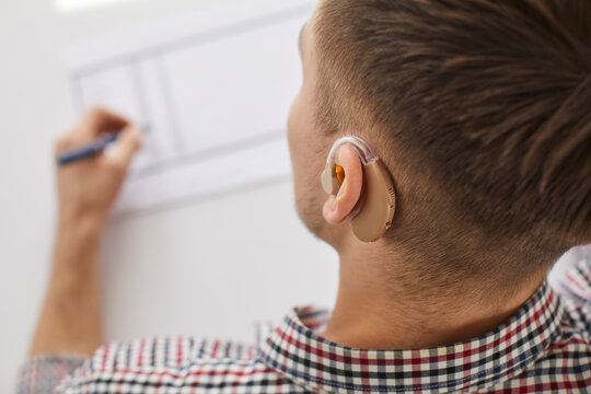 Close Up Shot Of Man With Hearing Aid Drawing On Paper. View From Behind Of Hearing Impaired Young Man Working At Desk In Office Or Home