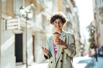 Obraz premium Portrait of a confident businesswoman enjoying a coffee break in the city. Young cheerful businesswoman walks outside with beaming smile while holding cup of coffee and laptop