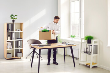 Fired male employee emptying his desk, making room for new worker. Young man standing by his former working desk in office and putting his belongings such as folders and green plants in cardboard box