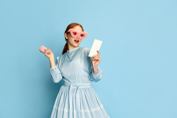 Portrait with young girl, princess with red hair wearing dress and pink plush sunglasses posing with an interested face over blue background. Concept of medieval era, beauty