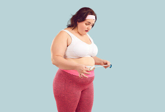 Woman finds out she has gained weight and has fat belly. Young overweight woman in sports bra and leggings looking at measuring tape that she holds around waist with surprised face expression
