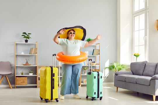 Full size photo of happy fat woman in big funny beach hat and sunglasses getting ready for holiday trip standing in the living room at home with two suitcases. Vacation and summer journey concept.