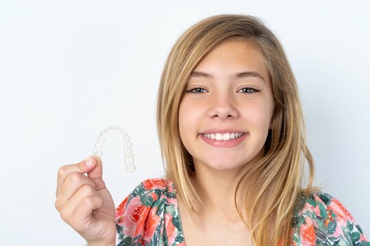 beautiful caucasian teen girl wearing flowered blouse over white wall holding an invisible braces aligner, recommending this new treatment. Dental healthcare concept.