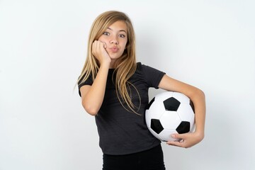 beautiful caucasian teen girl wearing sportswear holding a football ball over white wall with surprised expression keeps hands under chin keeps lips folded makes funny grimace