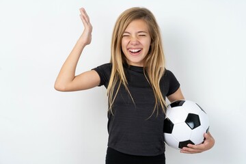 beautiful caucasian teen girl wearing sportswear holding a football ball over white wall goes crazy...