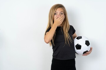 Stunned beautiful caucasian teen girl wearing sportswear holding a football ball over white wall...