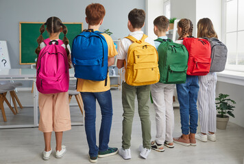 Back to school concept. Group of school children with backpacks. Backside view from behind six students with colorful pink, blue, yellow, green, red and grey bags standing together in the classroom