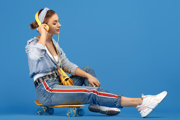 Full length photo of carefree young woman with yellow headphones sitting on skateboard over blue...