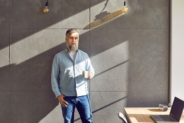 Portrait of a mature bearded attractive business man standing with a cup of tea or coffee near his workplace at office looking at the camera resting from a work on a grey wall background.