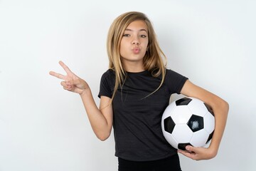 beautiful caucasian teen girl wearing sportswear and football ball over white wall makes peace gesture keeps lips folded shows v sign. Body language concept