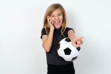 Positive beautiful caucasian teen girl wearing sportswear and football ball over white wal indicates directly at camera has telephone conversation smiles broadly enjoys talking long hours. You join me