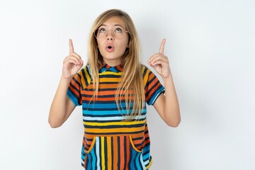 beautiful caucasian teen girl wearing striped T-shirt over white wall being amazed and surprised looking and pointing up with fingers showing something strange.