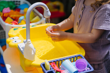 Child washes his hands and playing with toy kitchen outdoors in summer. Play area including running water. Developing Montessori toddlers activities. Kid play with water on fresh air