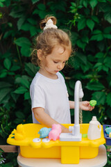 A little girl playing with toy kitchen in garden. Child play outdoors in summer. Play area including running water. Developing Montessori toddlers activities. Kid play with water on fresh air