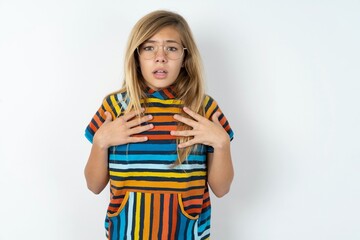 beautiful caucasian teen girl wearing striped T-shirt over white wall keeps hands on chest feeling shocked and scared, mouth widely opened, stares at camera saying: Who, me?