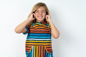 Fototapeta premium Stop making this annoying sound! Unhappy stressed out beautiful caucasian teen girl wearing striped T-shirt over white wall making worry face, plugging ears with fingers, irritated with loud noise.