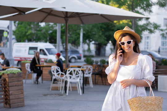 Young woman, a tourist in a white dress, straw hat, white glasses, straw bag, talking on the phone in the city, a cafe. Concept: the use of technology, smartphone, vacation, tourist in the city
