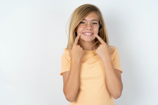 Strong healthy straight white teeth. Close up portrait of happy beautiful caucasian teen girl wearing orange T-shirt over white wall with beaming smile pointing on perfect clear white teeth.