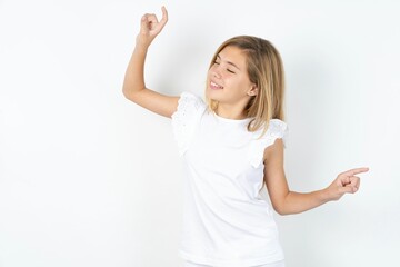 Photo of upbeat beautiful caucasian teen girl wearing white T-shirt over white wall has fun and dances carefree wear being in perfect mood makes movements. Spends free time on disco party